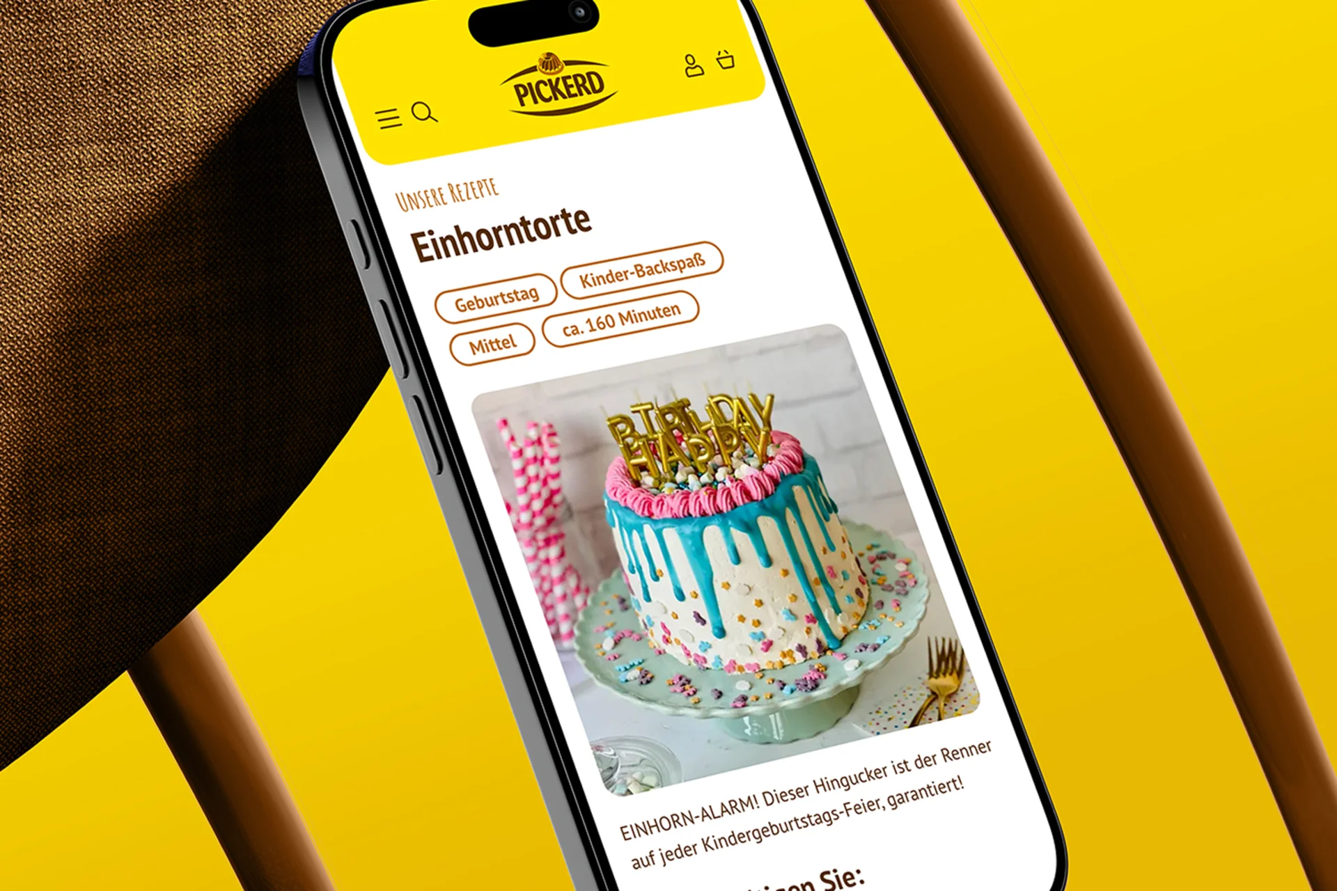 Smartphone vor braun, gelbem Hintergrund mit geöffneter Pickerd-Website zeigt Rezeptbild für eine Happy Birthday-Torte